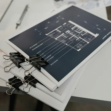 A stack of blueprints rests on a desk alongside a pen and scissors, indicating preparation for a design project.